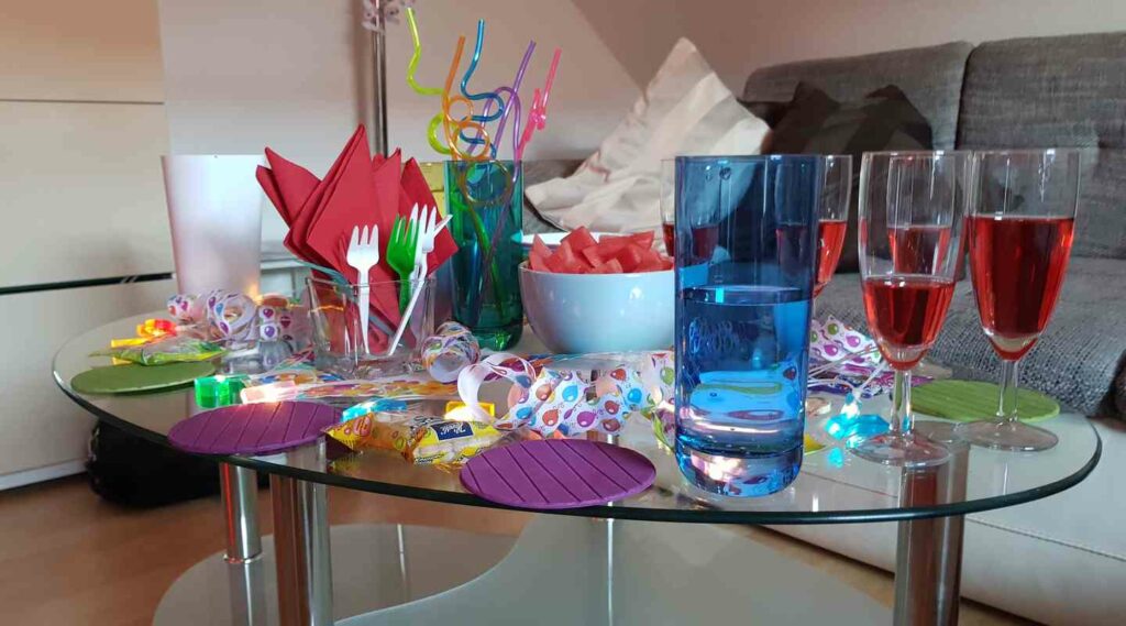 Glastisch mit bunten Strohhalmen, Tellern, roten Servietten, Gläsern und Snacks.