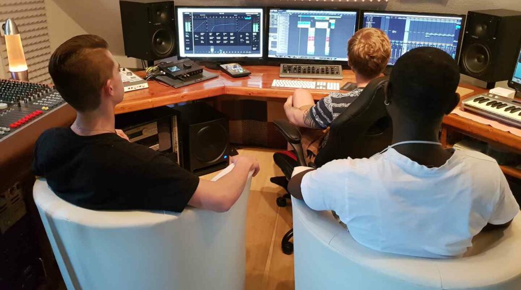 Drei Personen sitzen vor mehreren Bildschirmen und Audiotechnik im Tonstudio.