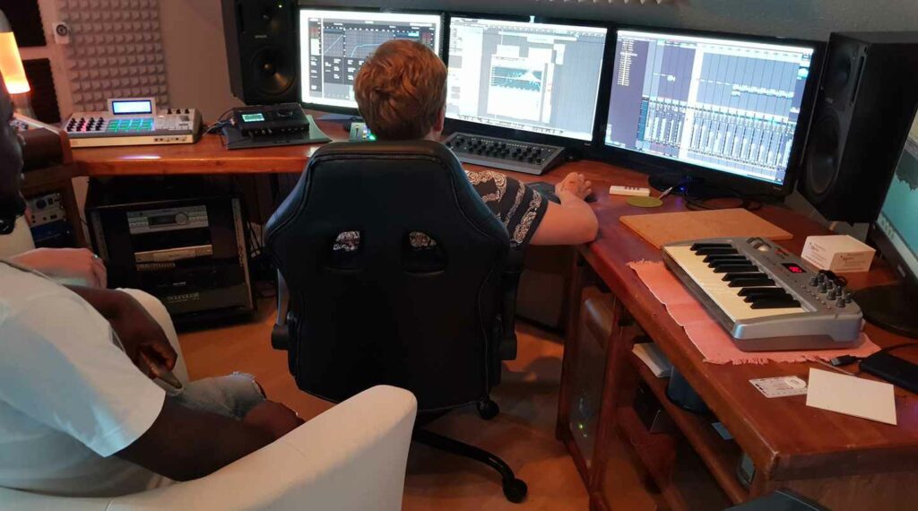 Eine Person bedient die Audioworkstation mit drei Bildschirmen; weitere Personen sitzen daneben.